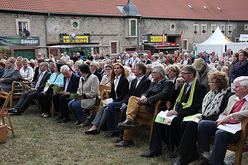 Viele Personen sitzen in Reihen auf Holzstühlen auf einer Wiese vor einem Podium.