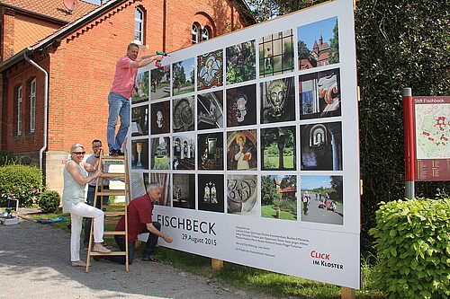 Vier Personen, eine davon auf einer Leiter, stehen vor einer Tafel, die mit Fotos bedruckt ist.