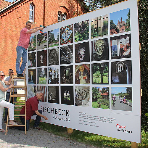 Vier Personen, eine davon auf einer Leiter, stehen vor einer Tafel, die mit Fotos bedruckt ist.
