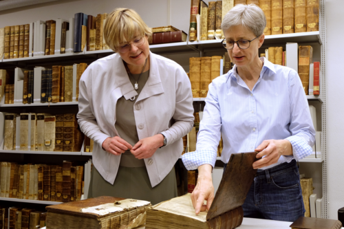 Zwei Frauen schauen sich historische Bücher an. Im Hintergrund ein Regal mit weiteren historischen Büchern.