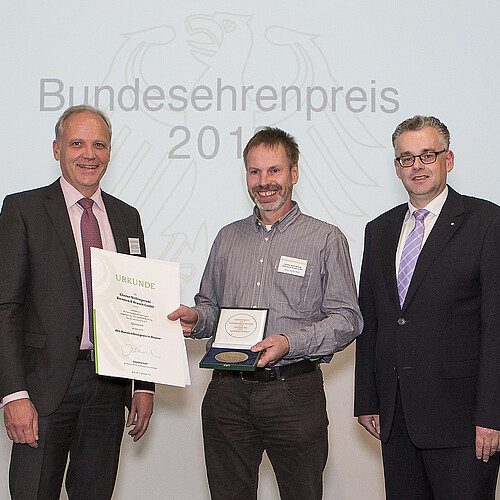 Drei Personen stehen nebeneinander: Der Mann in der Mitte hat eine Urkunde und eine Plakette in den Händen.