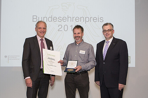 Drei Personen stehen nebeneinander: Der Mann in der Mitte hat eine Urkunde und eine Plakette in den Händen.