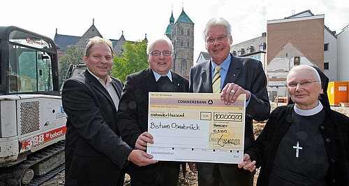 Vier Personen stehen auf einer Baustelle und halten gemeinsam einen übergroßen symbolischen Scheck.