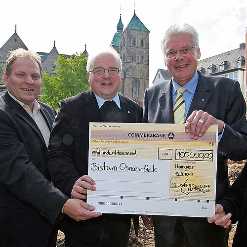 Vier Personen stehen auf einer Baustelle und halten gemeinsam einen übergroßen symbolischen Scheck.