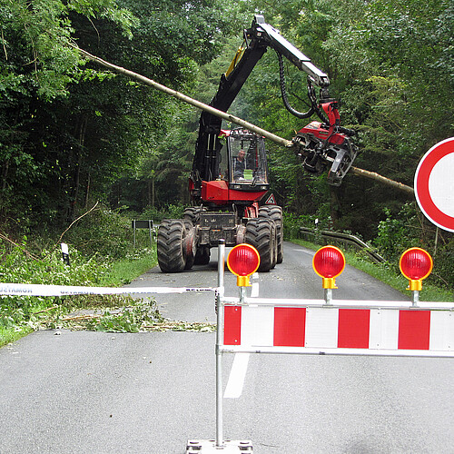 Im Vordergrund ist eine Straßensperrung zu sehen, im Hintergrund fährt eine Baumerntemaschine über die Straße.