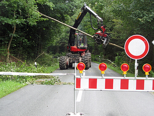 Im Vordergrund ist eine Straßensperrung zu sehen, im Hintergrund fährt eine Baumerntemaschine über die Straße.