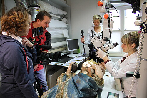 Eine Redakteurin, ein Kameramann und eine Tonfrau stehen um eine Restauratorin herum, die eine liegende Holzskulptur vor sich hat.