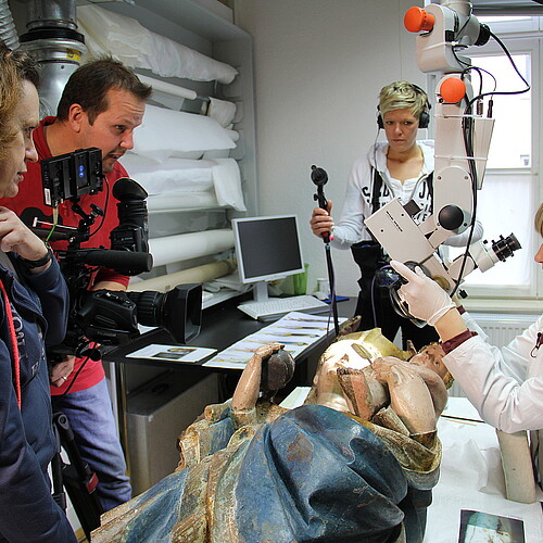 Eine Redakteurin, ein Kameramann und eine Tonfrau stehen um eine Restauratorin herum, die eine liegende Holzskulptur vor sich hat.