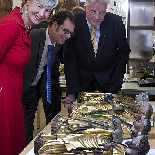 Drei Personen sind über teils vergoldete Figuren, die auf einem Tisch liegen, gebäeugt.