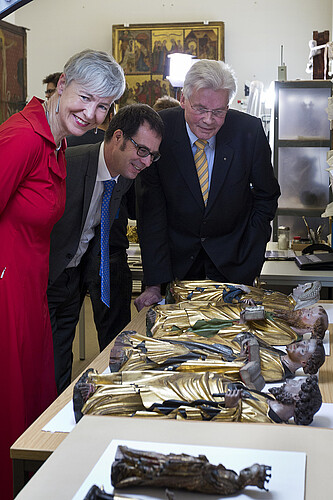 Drei Personen sind über teils vergoldete Figuren, die auf einem Tisch liegen, gebäeugt.