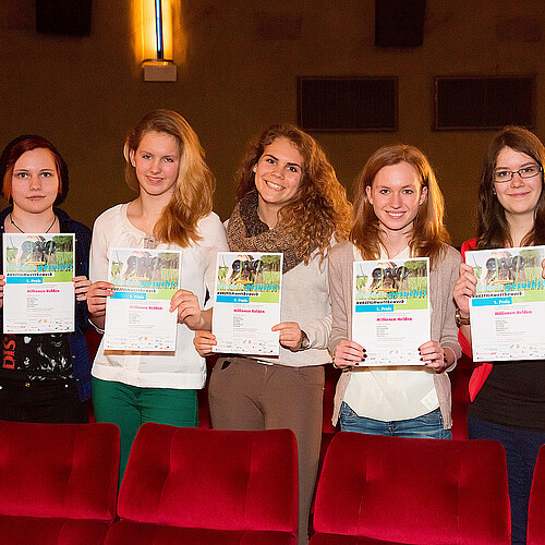 Fünf Schülerinnen, die jeweils eine Urkunde in den Händen halten, stehen zwischen Sitzreihen in einem Kino.