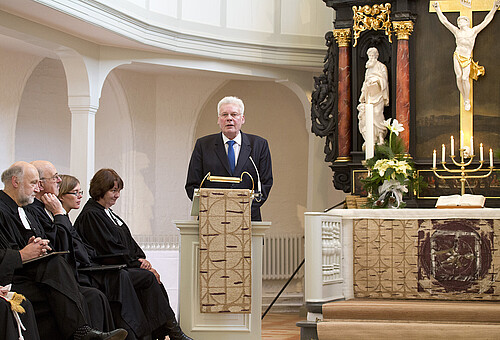 Klosterkammer-Präsident Hans-Christian Biallas steht hinter einem Rednerpult, neben einem Altar und einer Gruppe von mehreren Personen.
