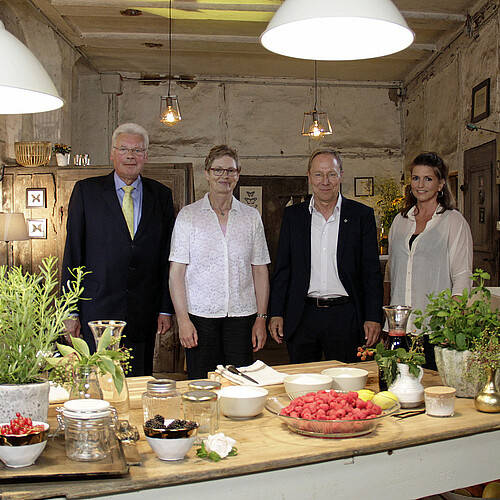 Vier Personen stehen in einer Küche, im Vordergrund ist ein Küchentisch mit Zutaten zu sehen.
