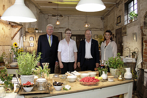 Vier Personen stehen in einer Küche, im Vordergrund ist ein Küchentisch mit Zutaten zu sehen.
