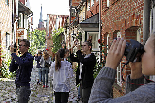 Mehrere Jugendliche stehen in einer Straße mit historischen Häusern und fotografieren.