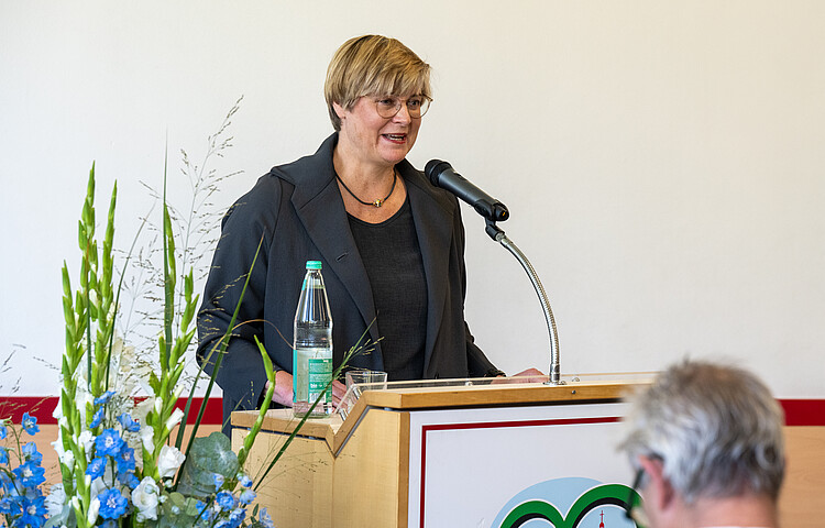 Eine Frau hält hinter einem Rednerpult stehend eine Rede. Vor ihr ein Mikrofon, links im Bild Blumenschmuck.