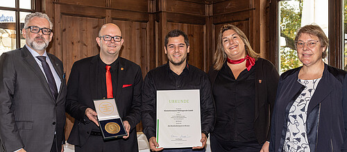 Drei Männer und zwei Frauen nebeneinander. Ein Mann hält Schachtel mit Medaille. Ein Mann hält eine Urkunde.