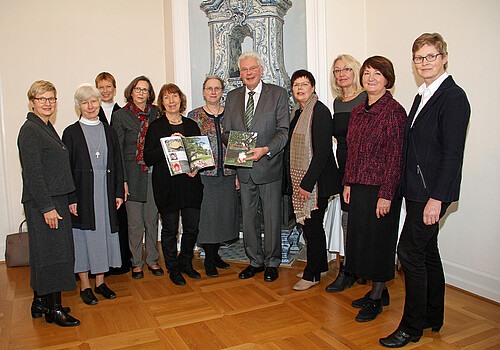 Zehn Frauen und ein Mann stehen in einem Halbkreis, zwei der Personen haben ein farbiges Magazin in der Hand.