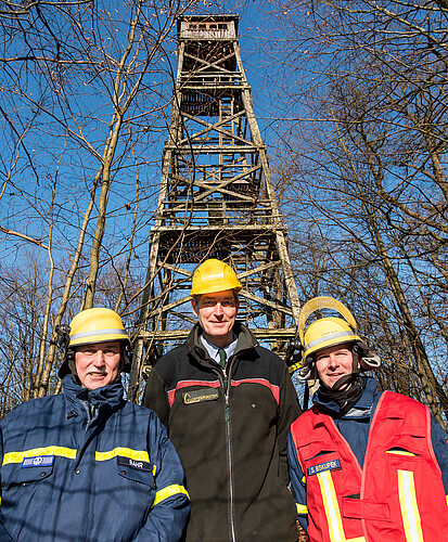 Drei Herren mit gelben Schutzhelmen stehen im Wald vor einem Turm aus hölzernen Streben.