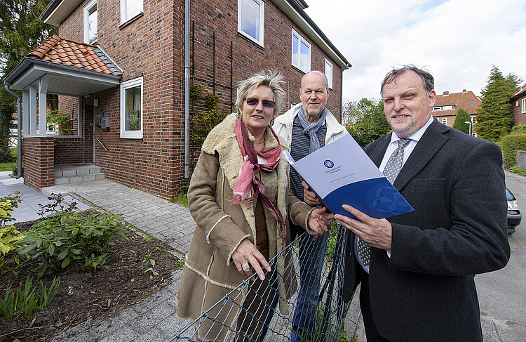 Eine Frau und zwei Männer stehen an einem Gartenzaun vor einem Haus beieinander. Der mann rechts ist Dieter Schulz, Leiter des Klosterrentamtes Lüneburg, hat eine Mappe in der Hand.