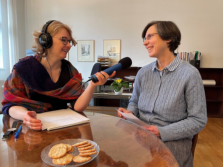 Zwei Frau an einem Tisch mit Keksschale, eine Frau mit Mikrofon und Kopfhörern interviewt die andere