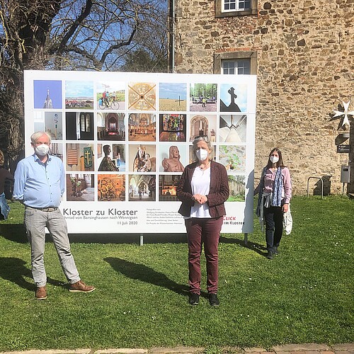 Zwei Frauen und ein Mann stehen jeweils mit Abstand zueinander vor einer großen Foto-Tafel vor dem Kloster Wennigsen.