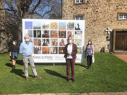 Zwei Frauen und ein Mann stehen jeweils mit Abstand zueinander vor einer großen Foto-Tafel vor dem Kloster Wennigsen.