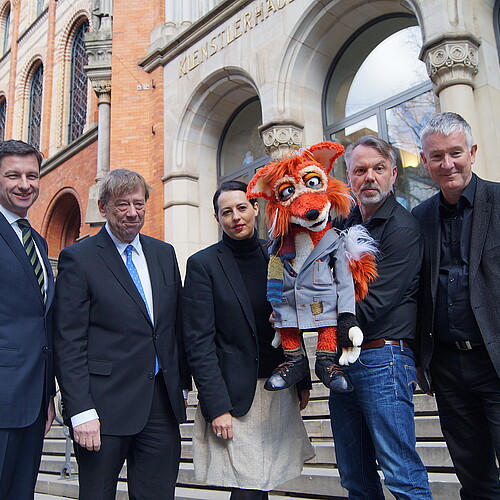 Fünf Personen, ein Mann hält eine Handpuppe, die einen Fuchs darstellt, stehen vor der historischen Fassade des Künstlerhauses in Hannover.