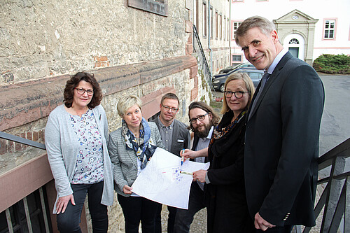 Sechs Personen, drei Frauen und drei Männer, stehen auf einer Treppe, die in ein Gebäude führt.
