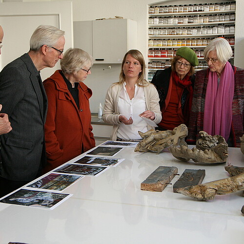 Fünf Personen hören Kirsten Schröder, Restauratorin in der Restaurierungswerkstatt der Klosterkammer Hannover, zu. Sie stehen dabei um einen Tisch in einer Werkstatt gruppiert.