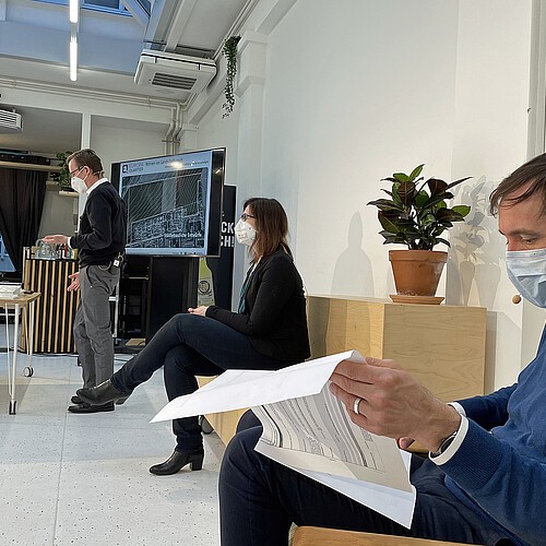 Eine Person steht vor einem Punkt und spricht, zwei andere sitzen jeweils mit Abstand, alle tragen Mund-Nase-Masen.