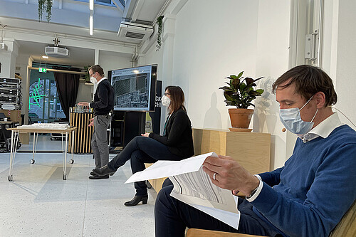 Eine Person steht vor einem Punkt und spricht, zwei andere sitzen jeweils mit Abstand, alle tragen Mund-Nase-Masen.