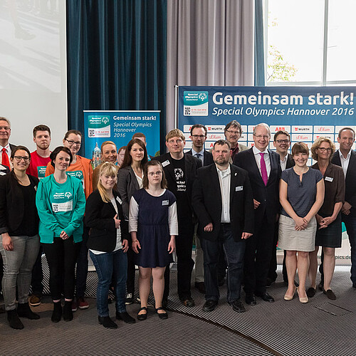 Eine große Gruppe von Personen mit und ohne Behinderungen steht gemeinsam in einem Saal.