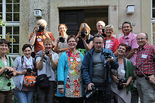 13 Personen stehen auf einer Treppe vor einem historischen Gebäude, sie halten bis auf Äbtissin Rosemarie Meding eine Kamera in der Hand.