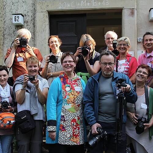 13 Personen stehen auf einer Treppe vor einem historischen Gebäude, sie halten bis auf Äbtissin Rosemarie Meding eine Kamera in der Hand.