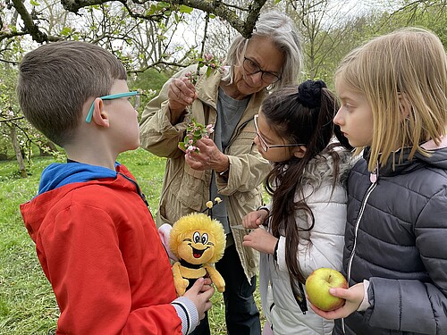Ältere Frau und drei Kinder stehen unter blühendem Apfelbaum.