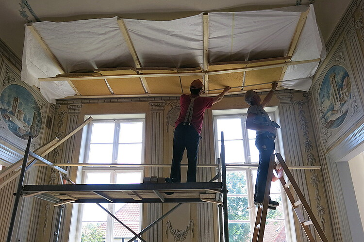 Zwei Männer stehen auf einen Gerüst und arbeiten an der Decke eines Raumes.