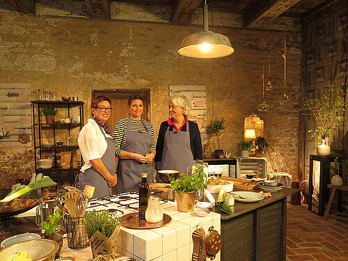 Drei Frauen stehen hinter einem hölzernen Küchentisch in einer historischen Küche.