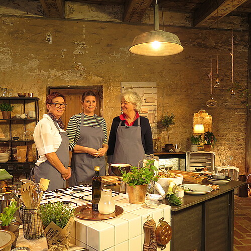 Drei Frauen stehen hinter einem hölzernen Küchentisch in einer historischen Küche.