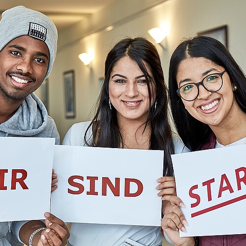 Drei Jugendliche halten je ein Blatt Papier in der Hand, darauf ist zu lesen: Wir sind START.