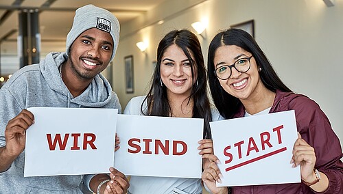 Drei Jugendliche halten je ein Blatt Papier in der Hand, darauf ist zu lesen: Wir sind START.