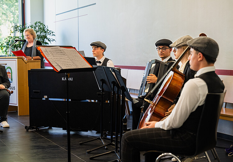 Vier junge Musiker unter anderem mit Klavier, Akkordeon und einem Streichinstrument sitzen auf einer Bühne. Im Hintergrund steht eine Frau an einem Rednerpult.