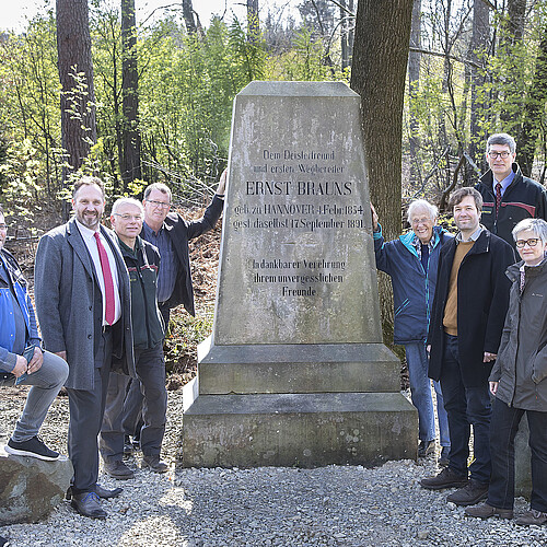 Acht Personen sind um ein steinernes Denkmal herum im Wald aufgestellt.