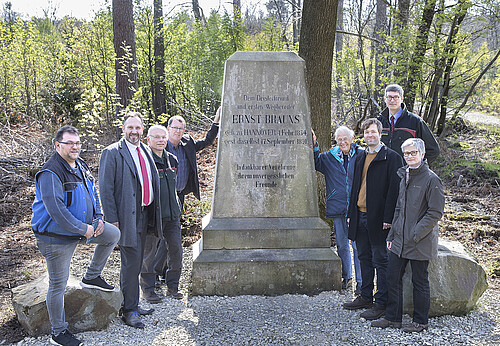 Acht Personen sind um ein steinernes Denkmal herum im Wald aufgestellt.