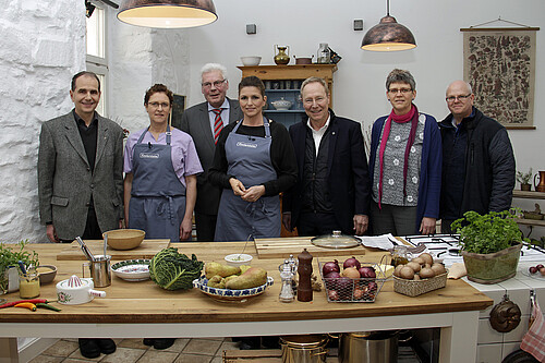 Drei Frauen und vier Männer stehen in einer für eine Fernsehaufzeichnung vorbereiteten Küche.