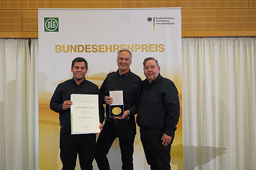 Drei schwarz gekleidete Männer mit einer Urkunde und einer Medaille