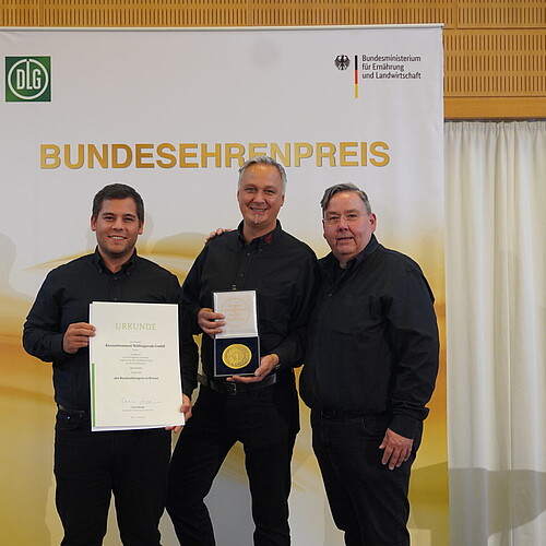 Drei schwarz gekleidete Männer mit einer Urkunde und einer Medaille