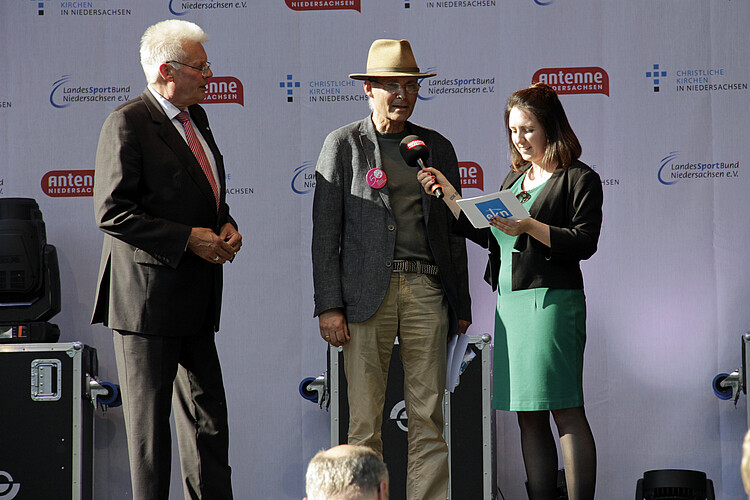 Zwei Männer und eine Frau stehen gemeinsam auf einer Bühne: Die Frau hält ein Mikrofon und Karten mit Notizen in der Hand.