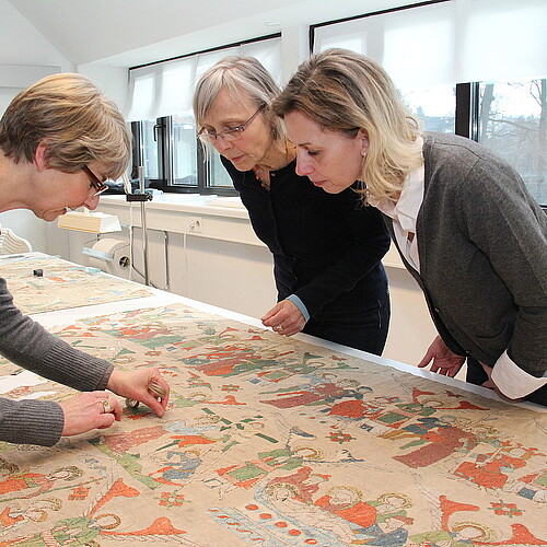 Drei Frauen betrachten ein mittelalterliches Altartuch in der Textilrestaurierungswerkstatt in Lüneburg.