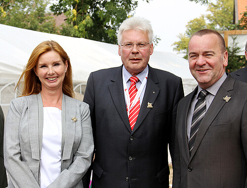 Eine Frau und zwei Männer stehen nebeneinander vor einem weißen Zelt im Freien.
